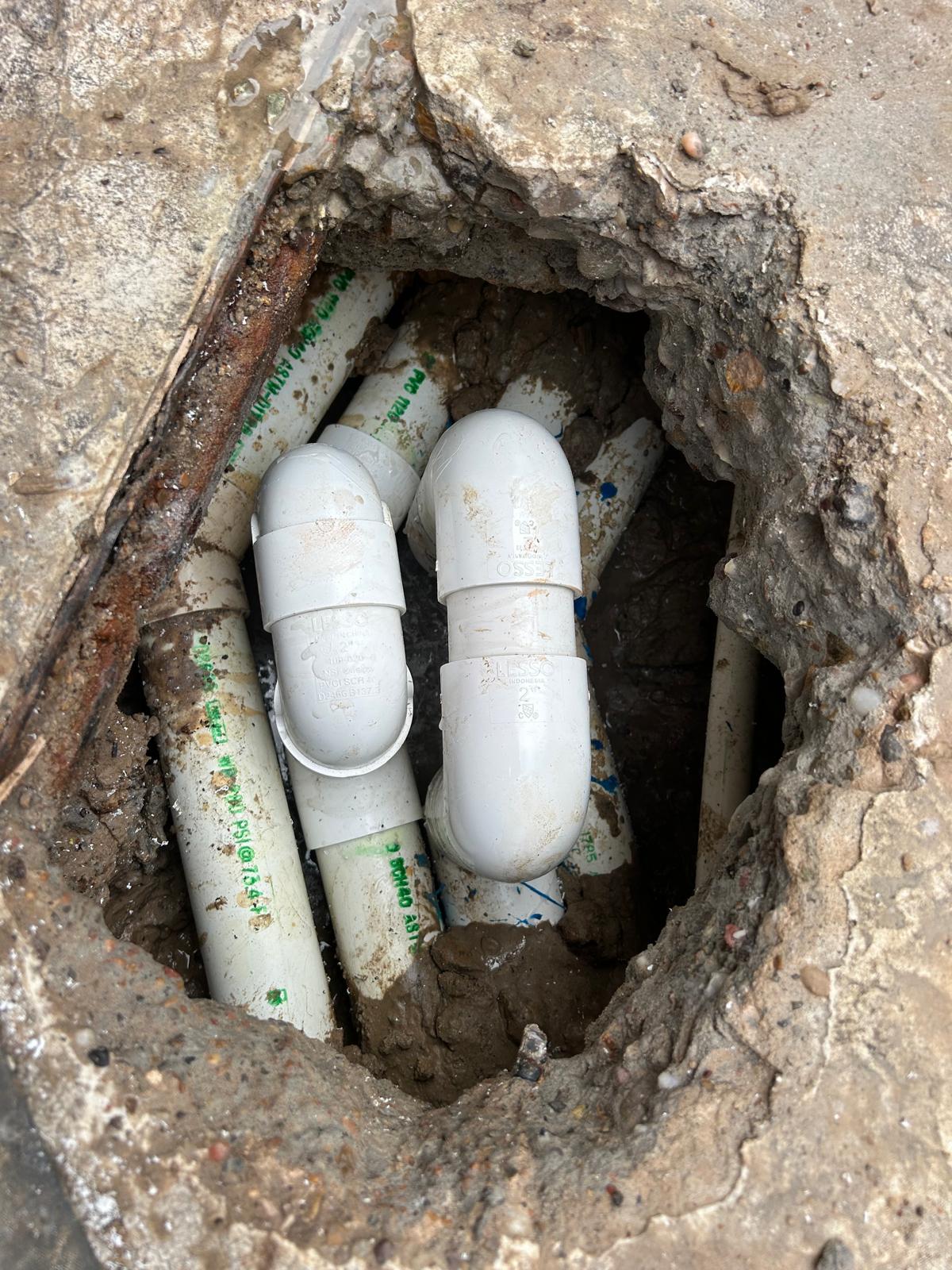 Small access hole cut through a concrete pool deck exposing a bundle of PVC pool lines with two new elbow repairs spliced in