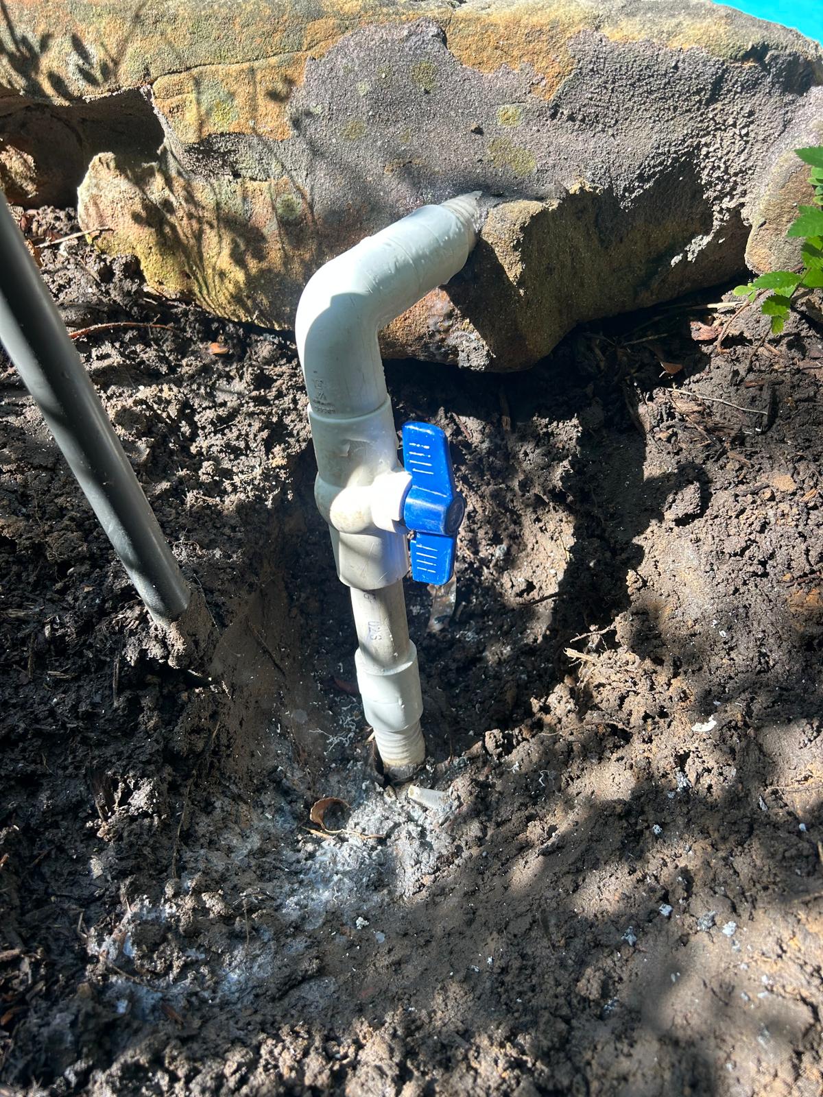 New blue handled PVC ball valve fitted into a pool water feature supply line in a small excavation beside the boulder coping